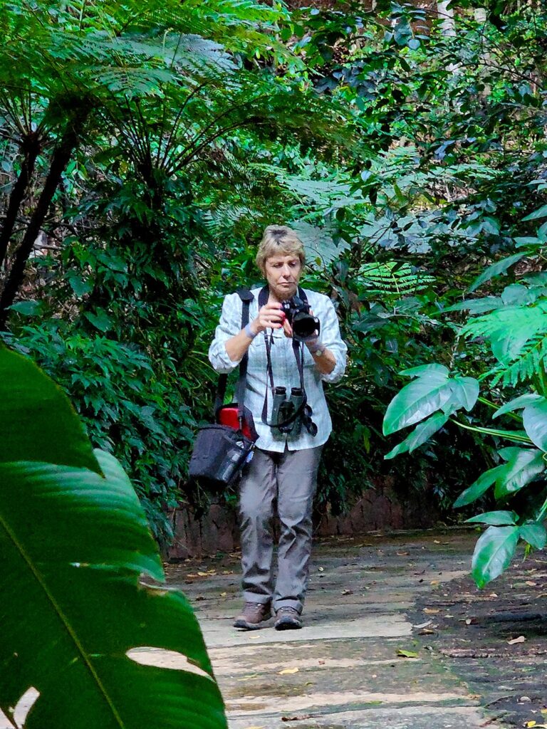 Foto mostrando a observadora Susanne Cohen enquanto ela tira uma foto. Ela segura uma câmera nas mãos e tem um binóculo preso ao pescoço por uma alça longa.