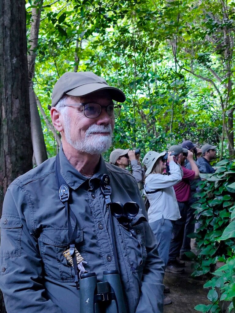 Foto apresentando o observador britânico John Reilly. Homem com cabelos brancos, óculos de grau e utilizando um boné e um casaco corta-vento verde escuro.