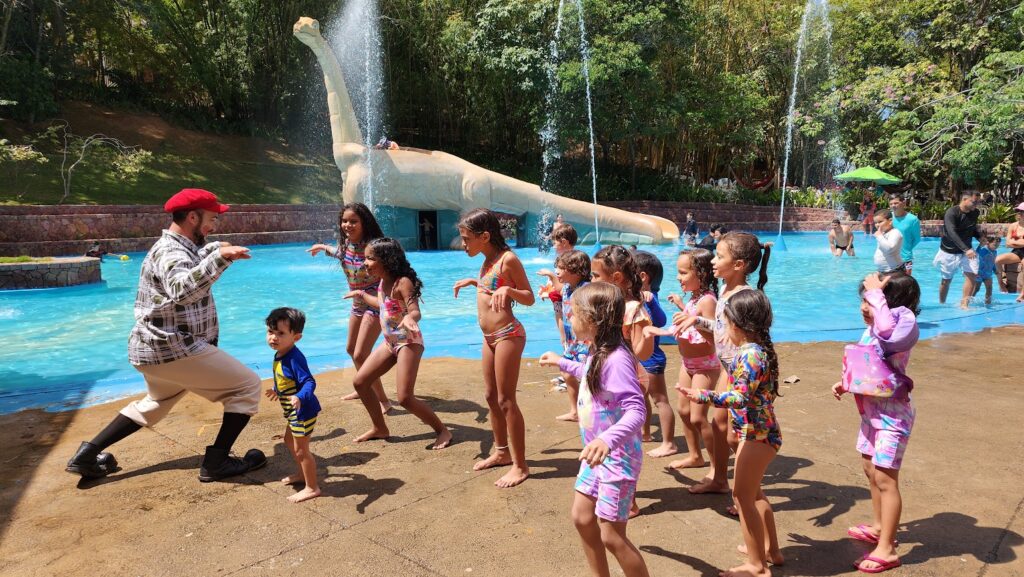 Crianças imitando um pássaro com o Palhação em um espaço aberto próximo a uma estátua de um Dinossauro.