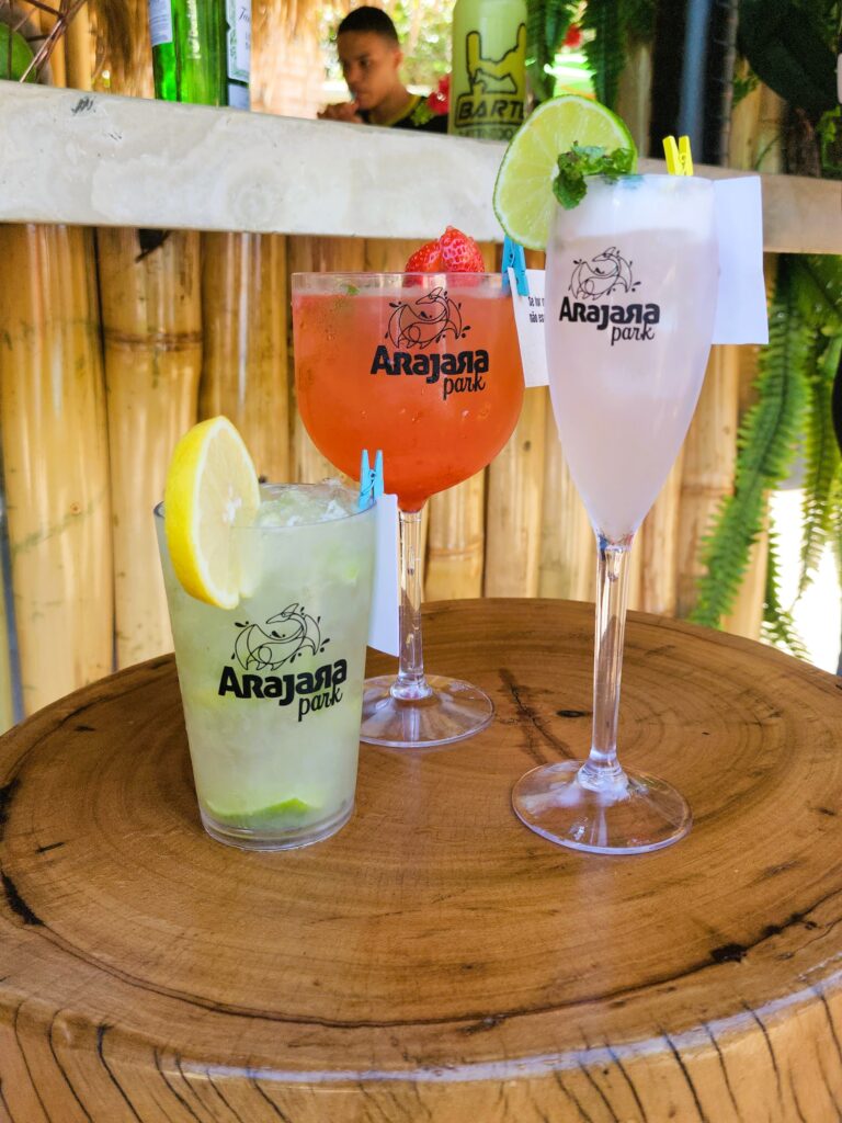 Foto em close de um copo e duas taças com bebidas. Ao fundo um bar de drinks.